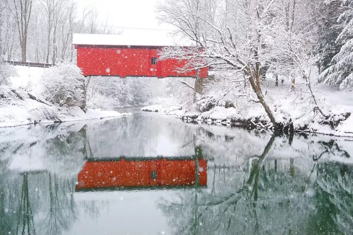 A red covered bridge reflected in a river, surrounded by snow-covered trees.